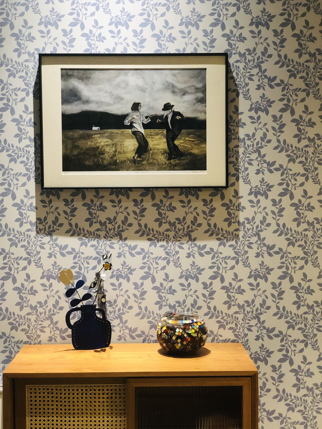 群青咖啡馆室内一角：蓝白叶纹壁纸、墙面挂着一幅油画，木质边柜上摆着蓝色陶壶与马赛克玻璃罐